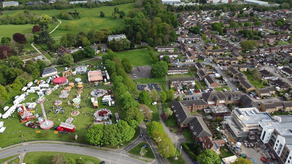 An aerial view of a park hosting a fair or community event, with various amusement rides including a large Ferris wheel, colorful tents, and food stalls. Surrounding the park are green trees, open gra